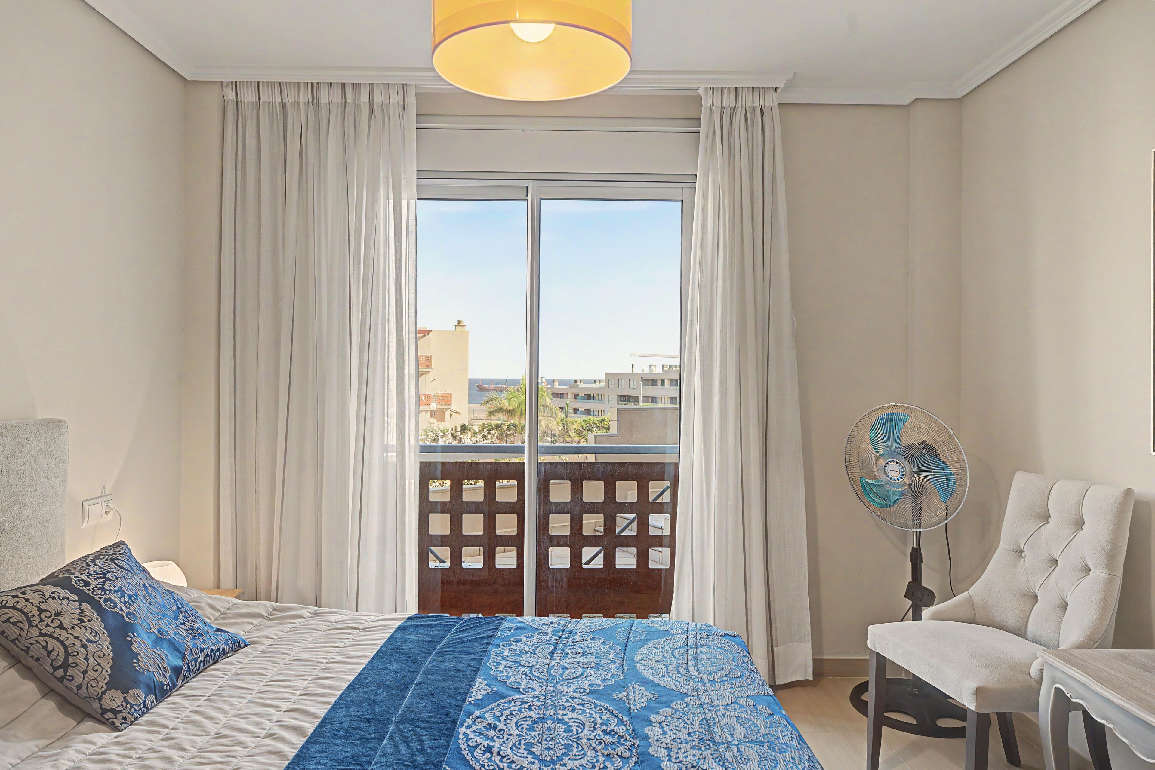 A bedroom with a bed, a chair, a fan, and a view of buildings through the window.