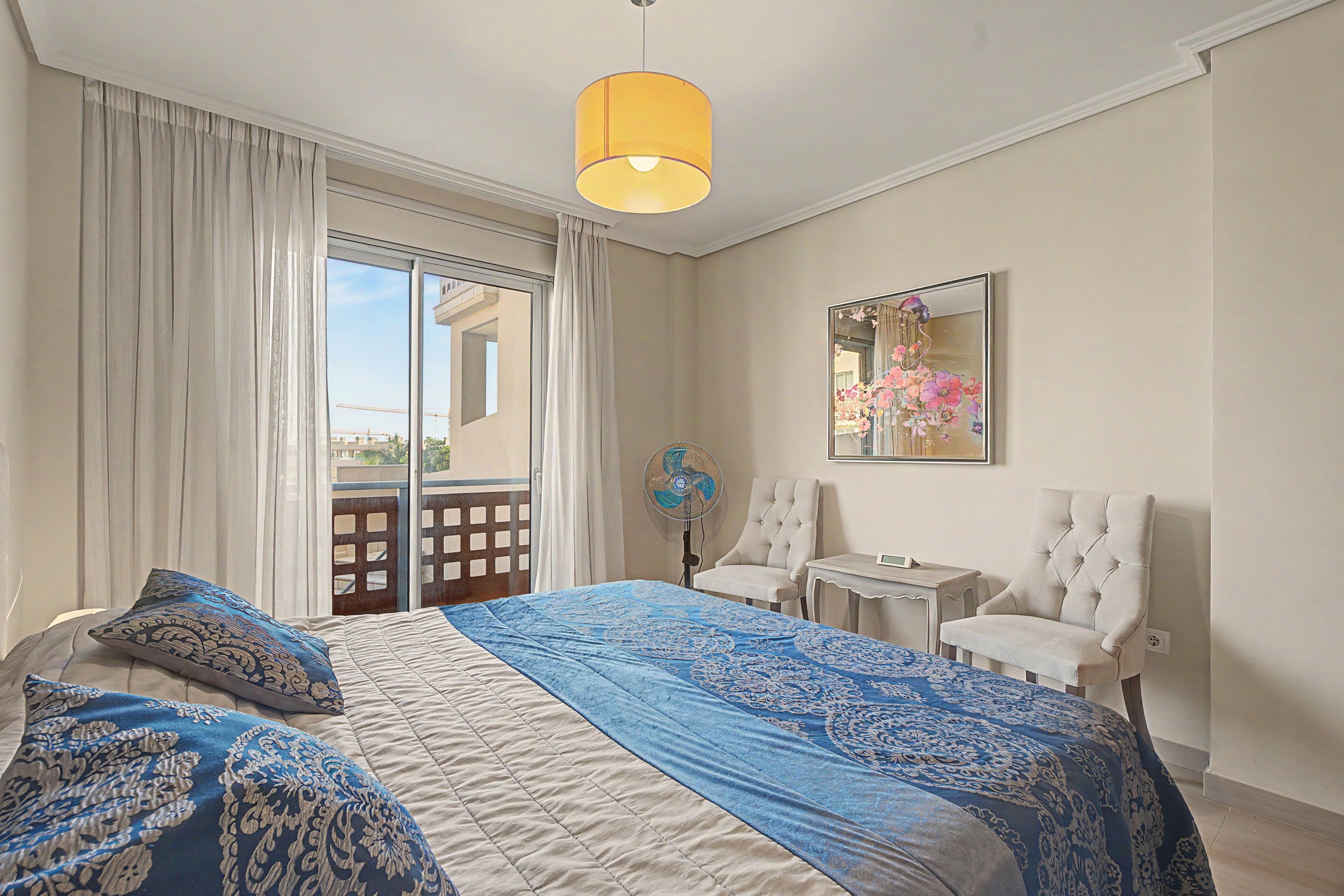 Ein Schlafzimmer mit einem Bett, einem Sessel, einem kleinen Tisch, einem Ventilator und einem Fenster mit Vorhängen.