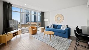 A modern living room with a large window, a blue sofa, a wooden coffee table, a TV, and a dining area. - Top Floor 2BR Penthouse - Great for Big Groups (Austin)