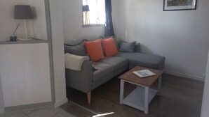 A modern living room with a grey sofa, orange cushions, a small wooden table, a lamp, and a framed picture on the wall. - Gite Barton 6 persons 500m from Utah Beach (Sainte-Marie-du-Mont)
