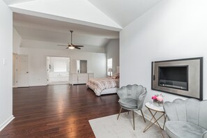 Ein geräumiger Raum mit Holzböden, einem Deckenventilator, einem Bett, einem grauen Sessel, einem kleinen Tisch mit einer Vase und einem an der Wand montierten Fernseher.