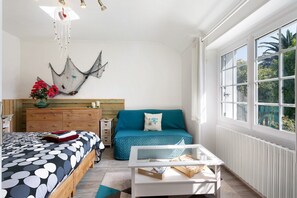 Ein Schlafzimmer mit Bett, Sofa, Couchtisch, Kommode und einem Fenster mit Blick ins Grüne.