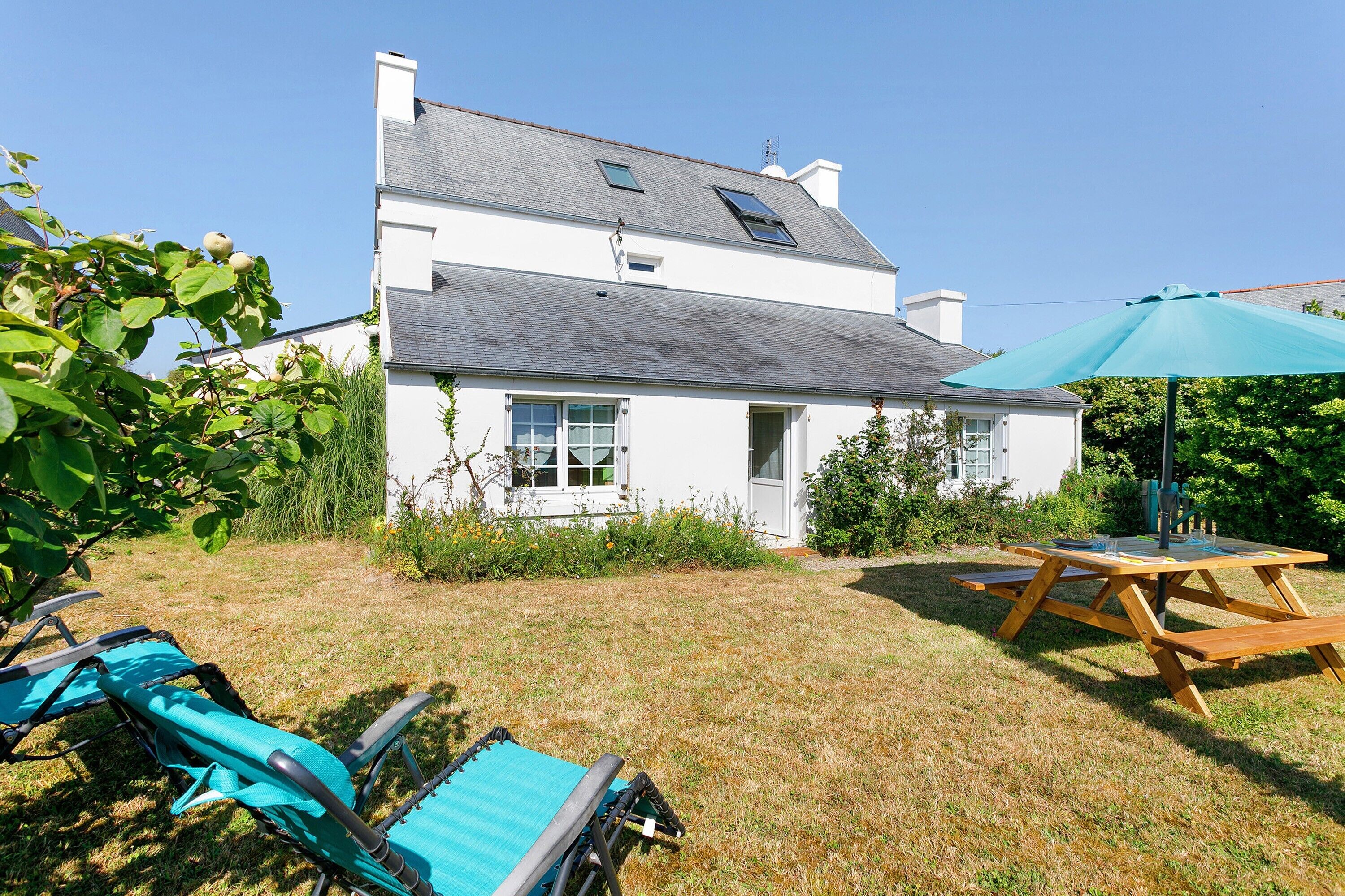 A white house with a sloped roof, a wooden picnic table, and two blue lawn chairs.