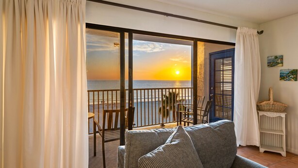 Ein Balkon mit Blick auf den Sonnenuntergang über dem Meer, eine Couch, Stühle und ein kleiner Tisch.