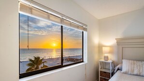 Ein Schlafzimmer mit großem Fenster und Blick auf einen Strand bei Sonnenuntergang, einem Nachttisch mit Lampe und einem ordentlich bezogenen Bett.