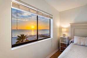 A bedroom with a large window overlooking a beach at sunset, a bedside table with a lamp, and a neatly made bed. - Beach Palms - Direct Gulf Front Condo (Indian Shores)