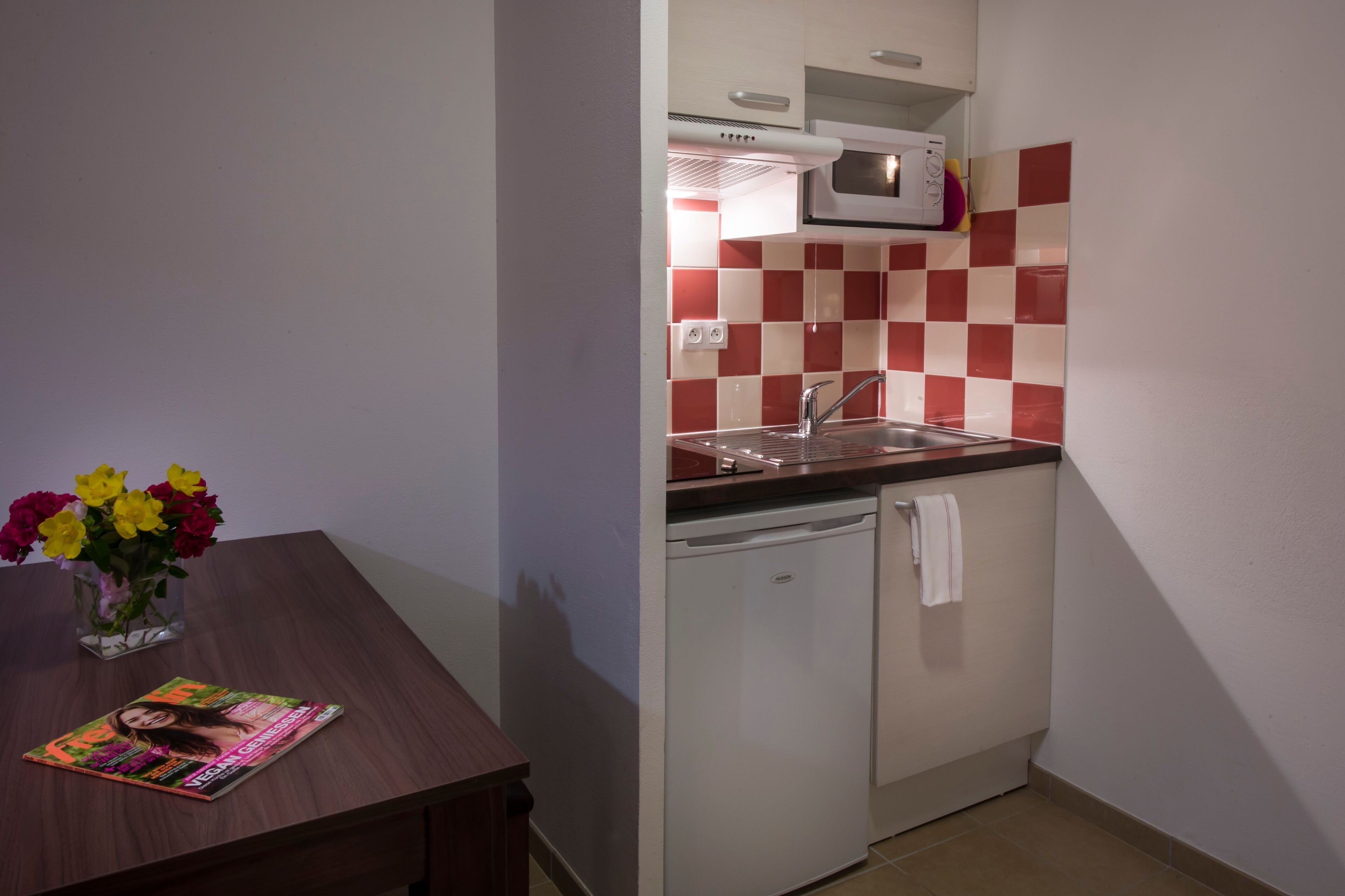 A compact kitchen with a microwave, sink, and dishwasher, a small table with a magazine and flowers, and a checkered backsplash.