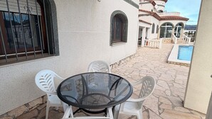 A patio area with a glass table and white plastic chairs, a stone floor, a pool, and a building with a tiled roof in the background. - Amarella Costa Dorada 1-bedroom apartment with pool (Les Tres Cales)