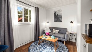 Een kamer met een grijze bank, een houten salontafel met een vaas en boeken, een rond blauw tapijt met patroon, een schilderij aan de muur en een tv op een houten dressoir.