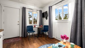Een kamer met een houten vloer, een ronde tafel met stoelen, een aan de muur bevestigde televisie en een raam met gordijnen.