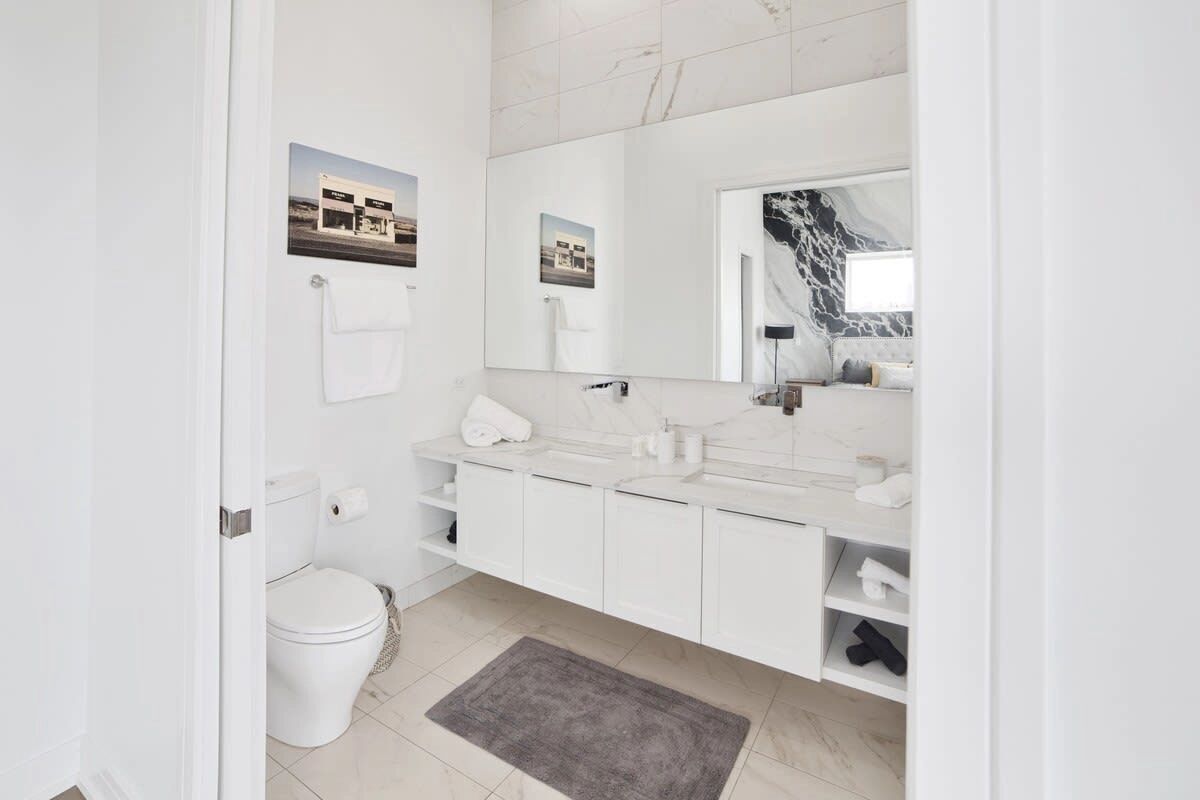 Een moderne badkamer met een toilet, twee wasbakken, een grote spiegel en een marmeren achterwand.
