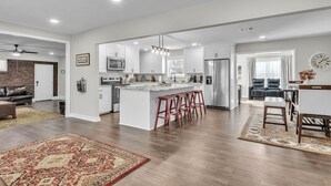 A modern kitchen with white cabinets, a central island with stools, a dining area with a table and chairs, and a living room with a sofa and a coffee table. - 4-bed/2-bath Spacious House in Charming Columbus, Game Day Home, Fenced Backyard (Columbus)