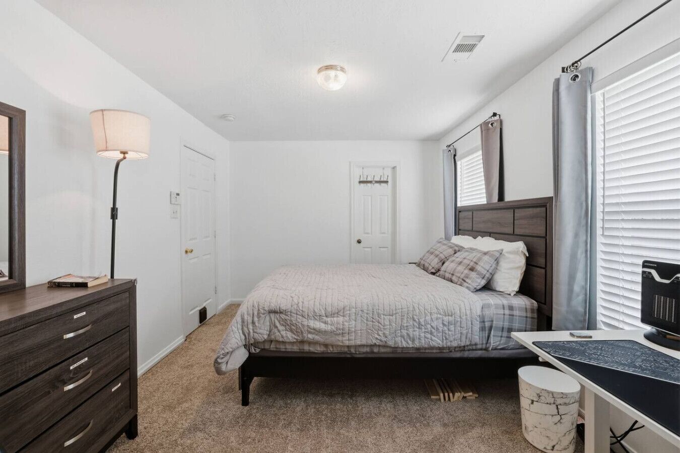 Een slaapkamer met een bed, een dressoir, een lamp, een bureau en een raam met jaloezieën.