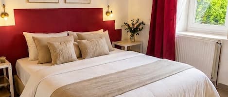 Een slaapkamer met een houten plafond, een groot bed met wit beddengoed, twee wandlampen en een raam met rode gordijnen.