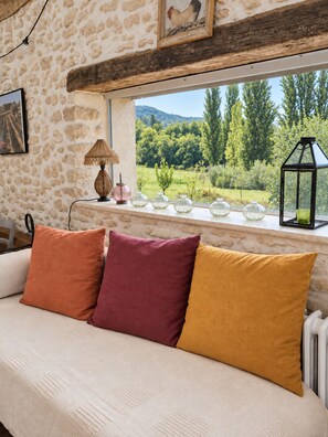 A room with a stone wall, a window with a view of greenery, a sofa with cushions, a lamp, and a lantern.