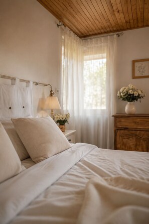 A bedroom with a wooden ceiling, a bed with white bedding, a nightstand with a lamp, a dresser, and a window with sheer curtains. - Le Mas d'Eylieux - Côté Source - Charm and views in southern Ardèche (Saint Montan)