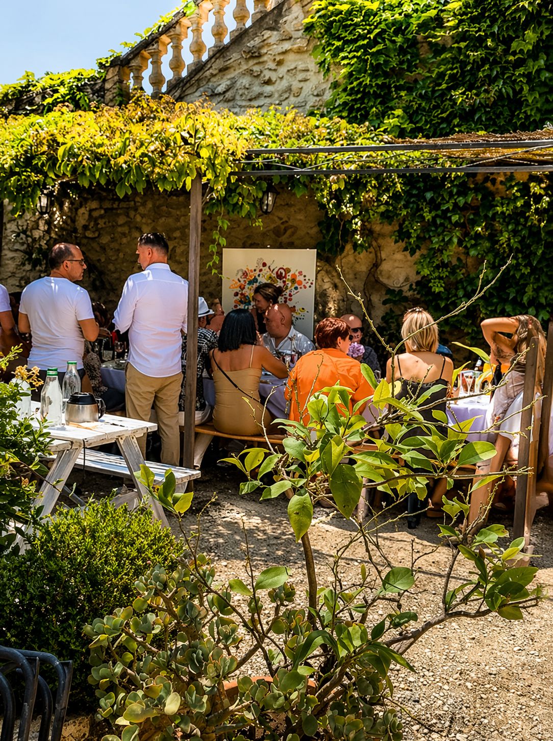 Une réunion en plein air où les participants sont assis à des tables, entourés de verdure et d’un mur de pierre.