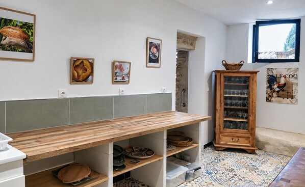 Une cuisine avec une table en bois, des étagères remplies d’ustensiles de cuisine, un meuble contenant de la vaisselle et des œuvres d’art mural.