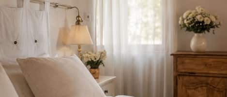 Een slaapkamer met een houten plafond, een bed met witte beddengoed, een nachtkastje met een lamp, een dressoir en een raam met dunne gordijnen.