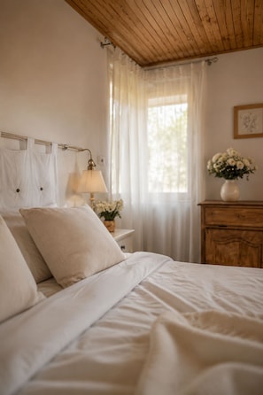 Un dormitori amb sostre de fusta, un llit amb roba de llit blanca, una tauleta de nit amb una làmpada, un armari i una finestra amb cortines transparents.
