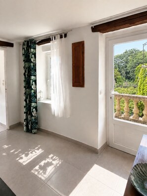 Una habitació amb un balcó, una finestra amb cortines blanques, una porta de fusta i vista a vegetació a l'exterior.
