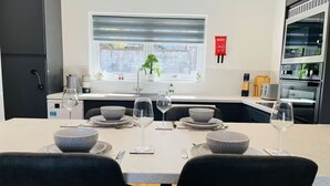 Uma sala de jantar com mesa posta para dois, uma janela com persianas, área de cozinha com eletrodomésticos e uma pequena planta no parapeito.