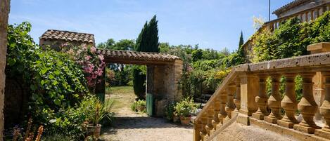 A garden pathway leading to a stone building with a tiled roof, surrounded by lush greenery and flowering plants.