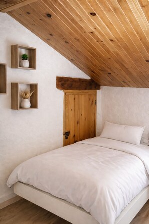 A bedroom with a sloped ceiling, wooden beams, a bed with white bedding, and two wall-mounted shelves with plants. - Châteaunette · Mezzanine and Pool (Saint Montan)
