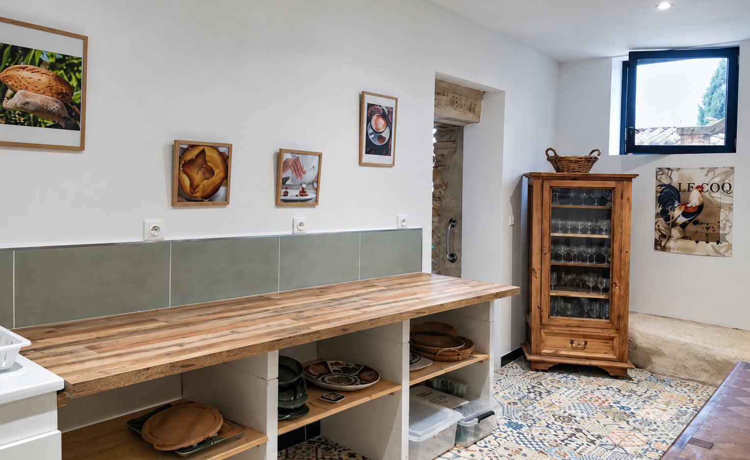 Een keuken met een houten tafel, planken met keukengerei, een kast met glazen en wanddecoratie.