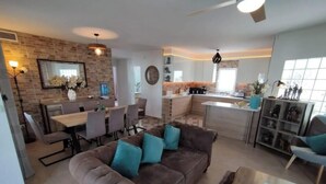 A modern living room with a brown sofa, dining area, kitchen, and a large window.