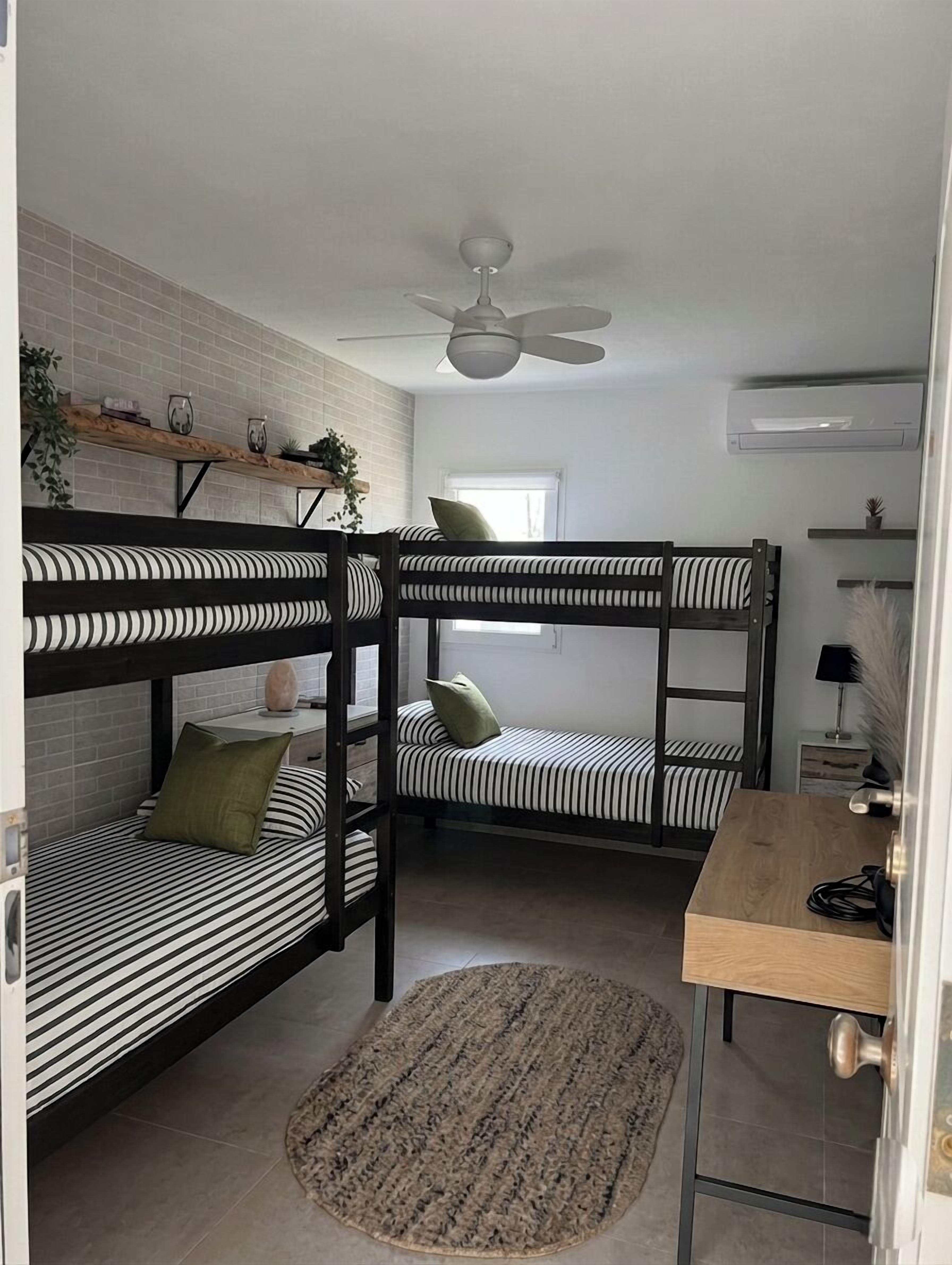 Ein Schlafsaalzimmer mit Etagenbetten, einem Deckenventilator, einem Schreibtisch und einem kleinen Fenster.