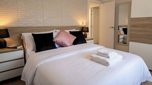 A neatly made bed with white bedding, black and pink pillows, and a stack of white towels. There are two bedside lamps on wooden nightstands.