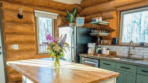 Eine Küche mit Holzwänden, einer zentralen Kochinsel mit Barhocker, einem Kühlschrank und einem Fenster mit Blick auf Bäume.