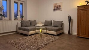 Ein Wohnzimmer mit einer Couch, einem Glastisch, einem Teppich, einem Schrank und einem Fenster mit Pflanzen.