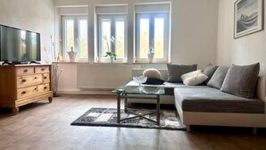 Ein Wohnzimmer mit einer Couch, einem Glastisch, einer Holzkommode, einem Fernseher und drei Fenstern.