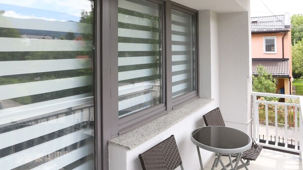 Un balcón con una mesa y sillas, ventanas amplias con persianas y vista a una zona residencial.