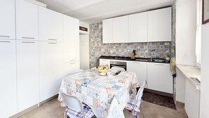 A compact kitchen with white cabinets, a tiled backsplash, a dining table with a patterned tablecloth, and a small island with a fruit bowl. - P092 – Porto Recanati, three-room apartment  (Porto Recanati)