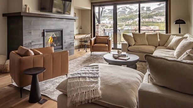A modern living room with a fireplace, leather sofas, a coffee table, and a large window with a mountain view. - New Build Condo with Shared Hot Tub (Big Sky)