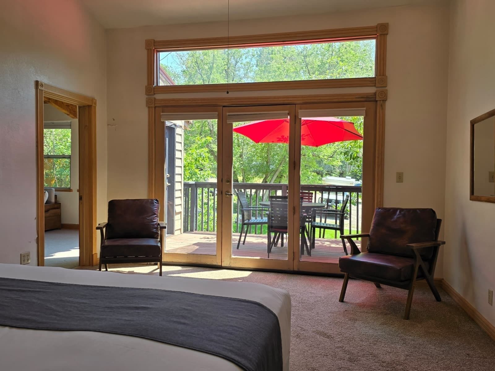 A bedroom with a large bed, a sliding glass door leading to an outdoor patio with a red umbrella, and two chairs.