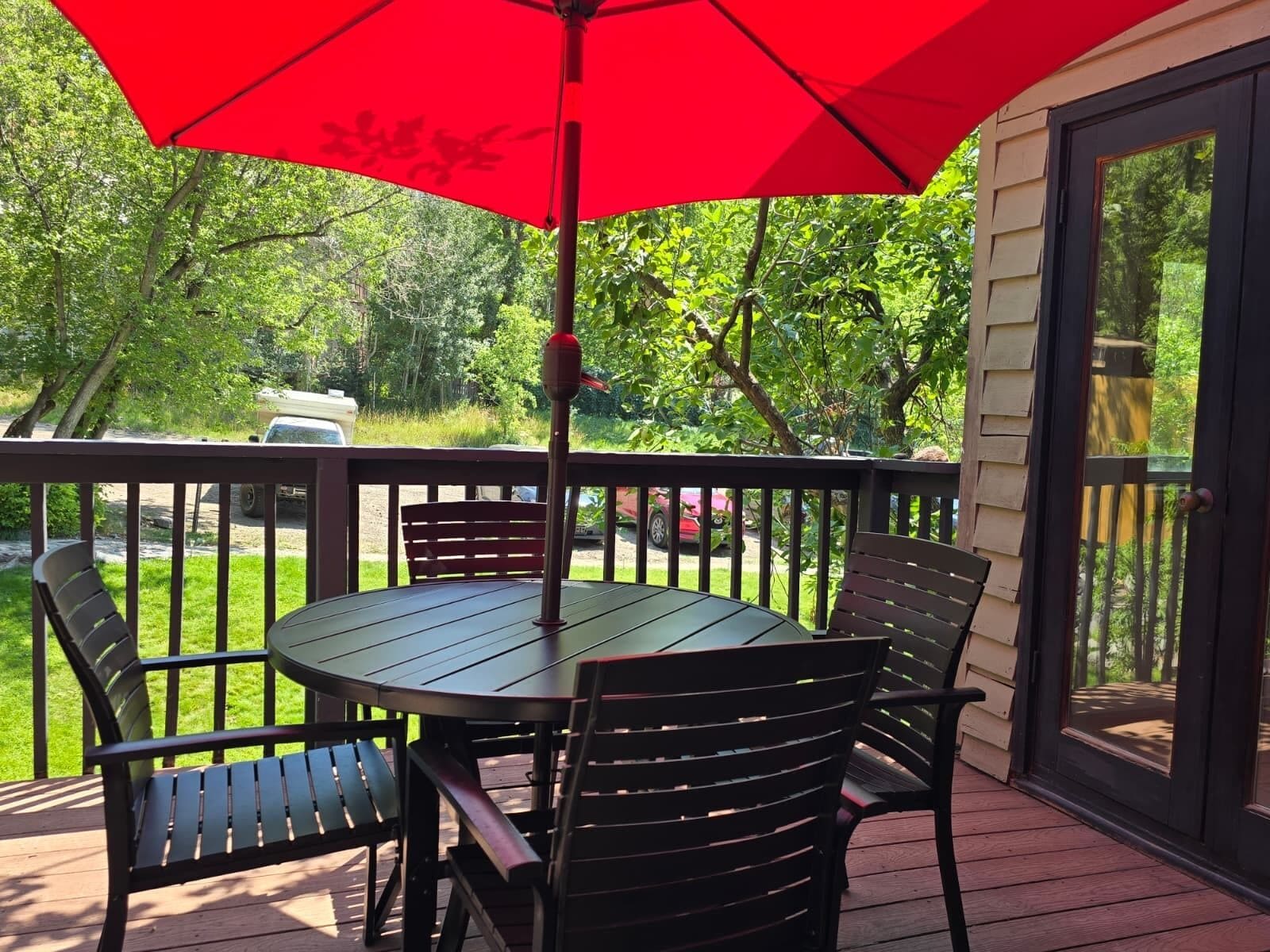 A covered outdoor seating area with a table and chairs, a red umbrella, and a view of a green landscape.