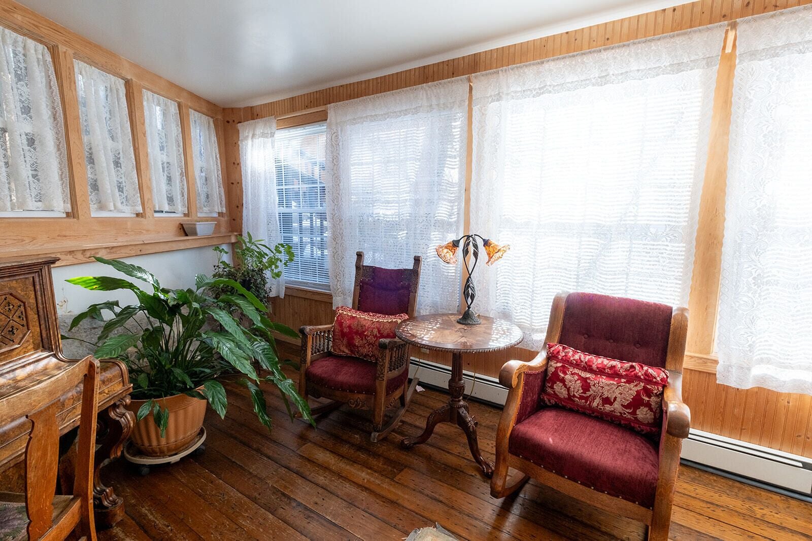 A room with wooden flooring, a wooden bench, two chairs, a table with a lamp, and a potted plant.