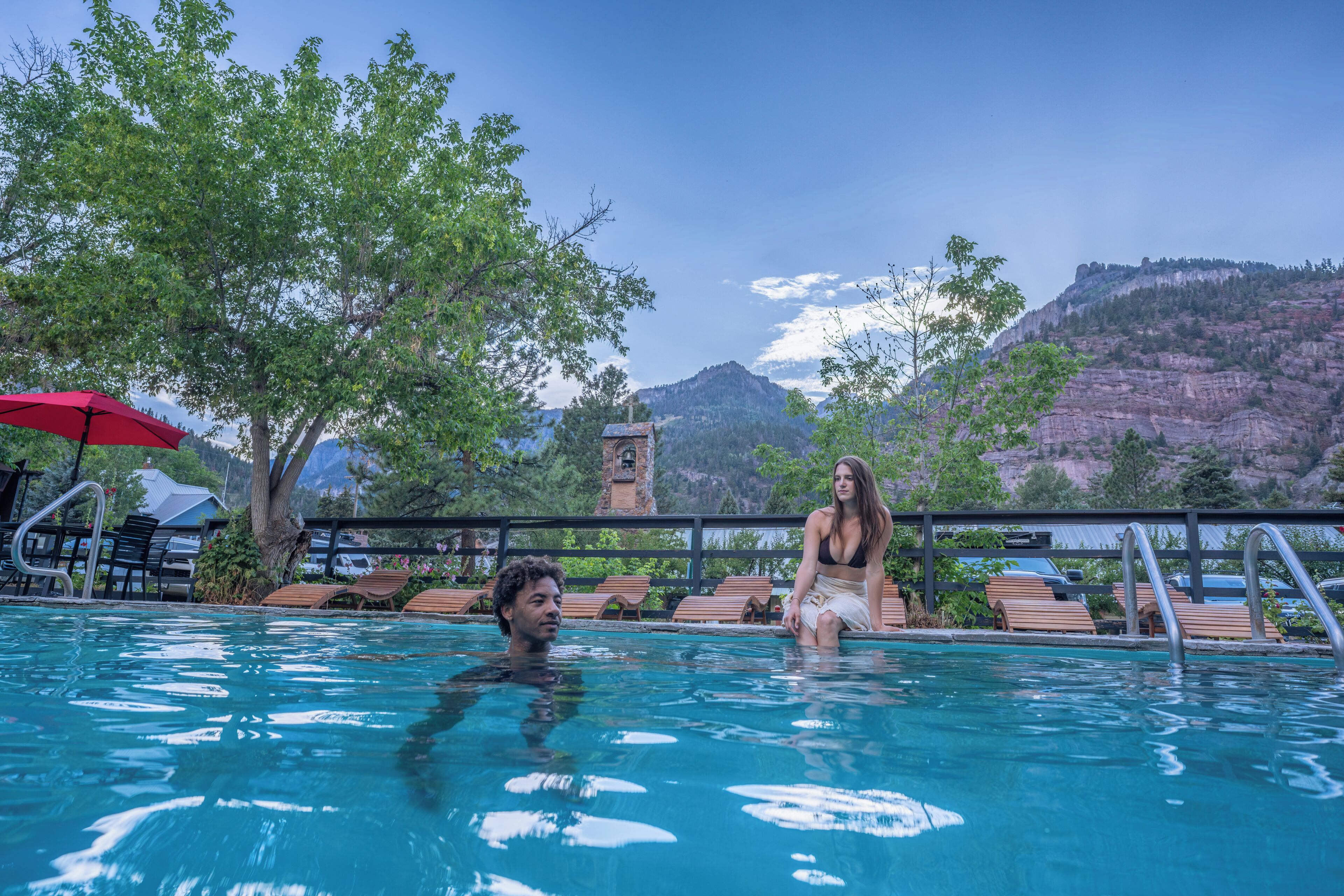 Una piscina con sillones de madera, una sombrilla roja y montañas al fondo.