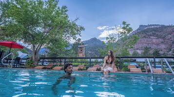 Una piscina con sillones de madera, una sombrilla roja y montañas al fondo.