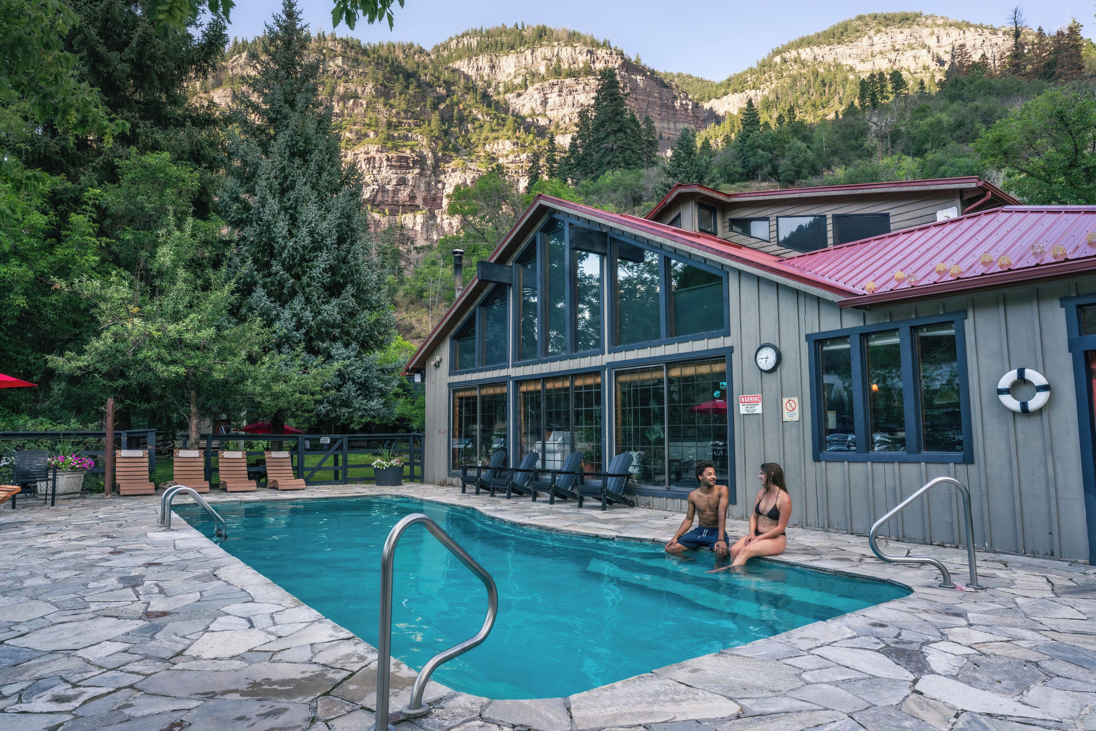 Un espace piscine doté d’une terrasse en pierre, de chaises longues et d’un bâtiment avec de grandes fenêtres et un toit rouge.