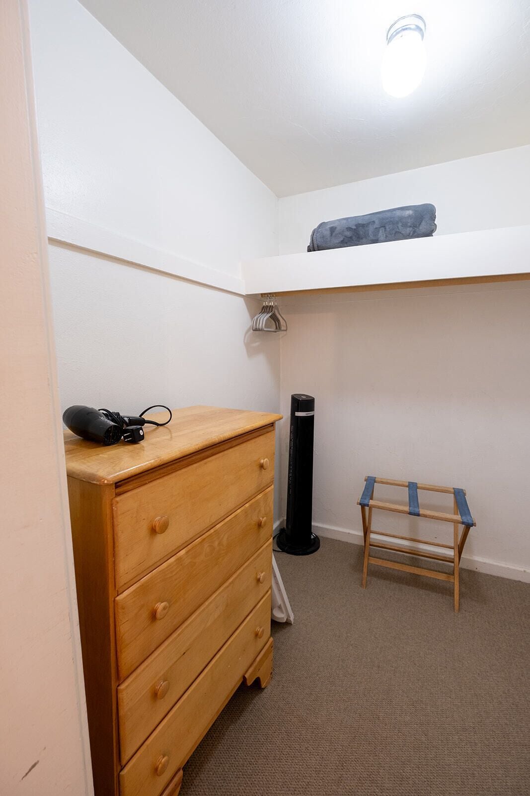 A small, well-lit room with a wooden dresser, a black standing heater, a wooden ladder, and a shelf with a rolled-up item.