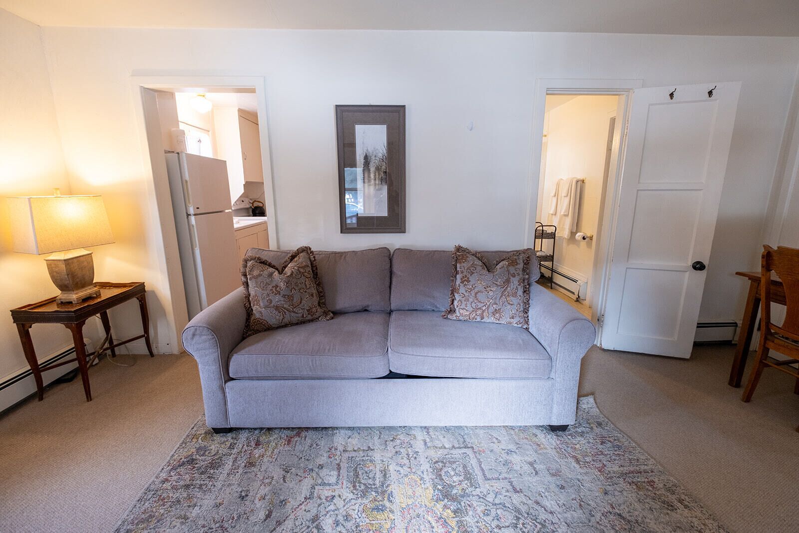 Una sala de estar con un sofá gris, alfombra estampada, una mesita con lámpara y una puerta que da a otra habitación.