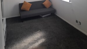 Une pièce avec un canapé gris, deux coussins orange, un mur blanc, une fenêtre et un luminaire suspendu.