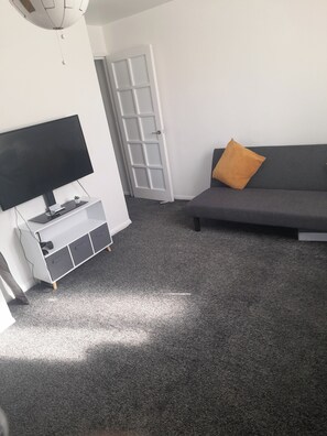 Un salon avec un téléviseur à écran plat posé sur un meuble blanc, un canapé gris agrémenté d’un coussin orange et une porte donnant sur une autre pièce.