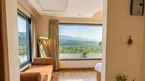 Una habitación con suelo de madera, un sofá marrón, una cama, un ventanal con vistas a zonas verdes y una planta en maceta.