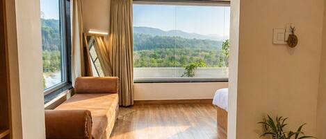 A room with a wooden floor, a brown sofa, a bed, a large window with a view of greenery, and a potted plant.
